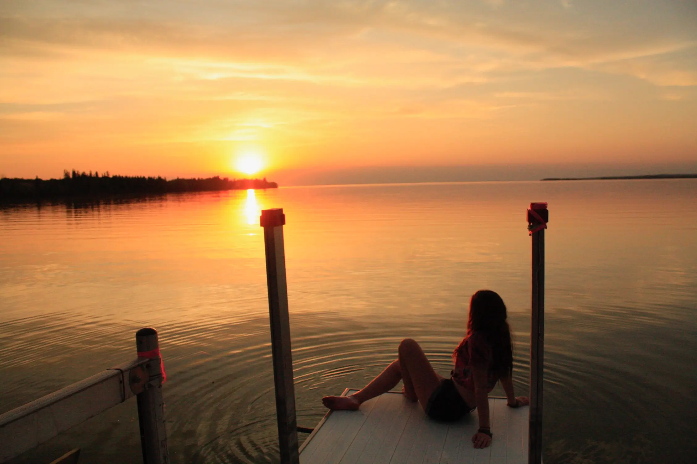 Sunset over Candle Lake with a person seated on a dock, reflected in still evening water.