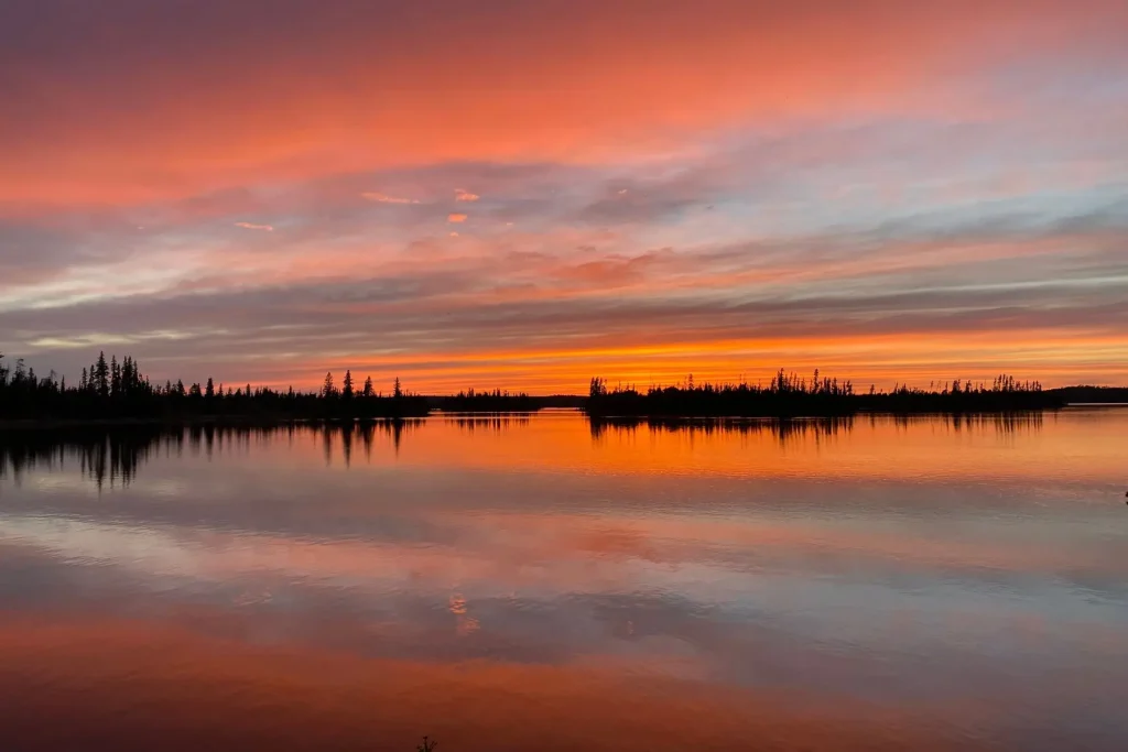 Fiery sunset reflecting on calm water along the Churchill River with silhouetted trees on the horizon.