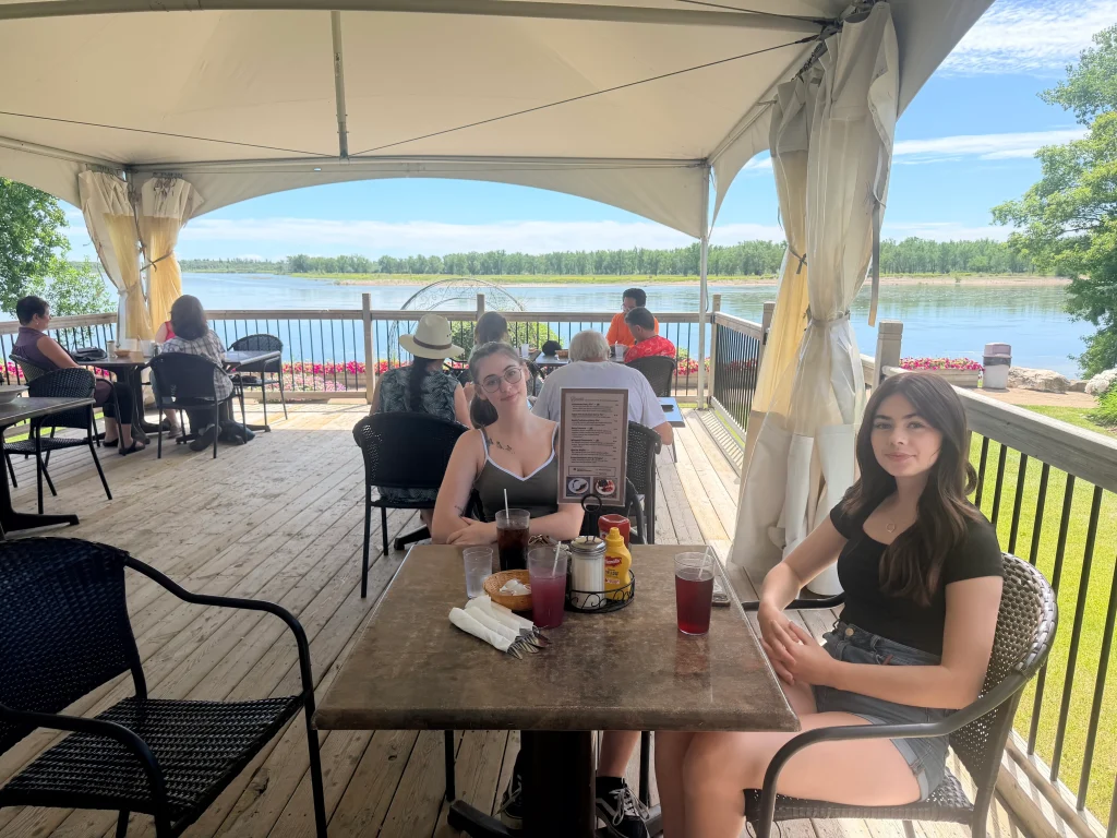 diners at a table on an outdoor patio with a view of a river and flower gardens in the background.