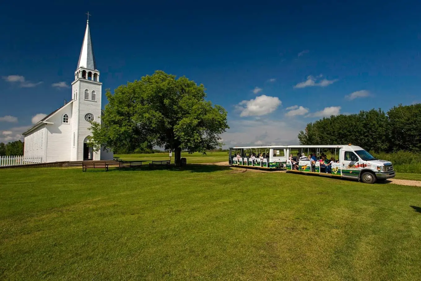 White church at Batoche National Historic Site with a tour tram carrying visitors across open prairie under a blue sky.