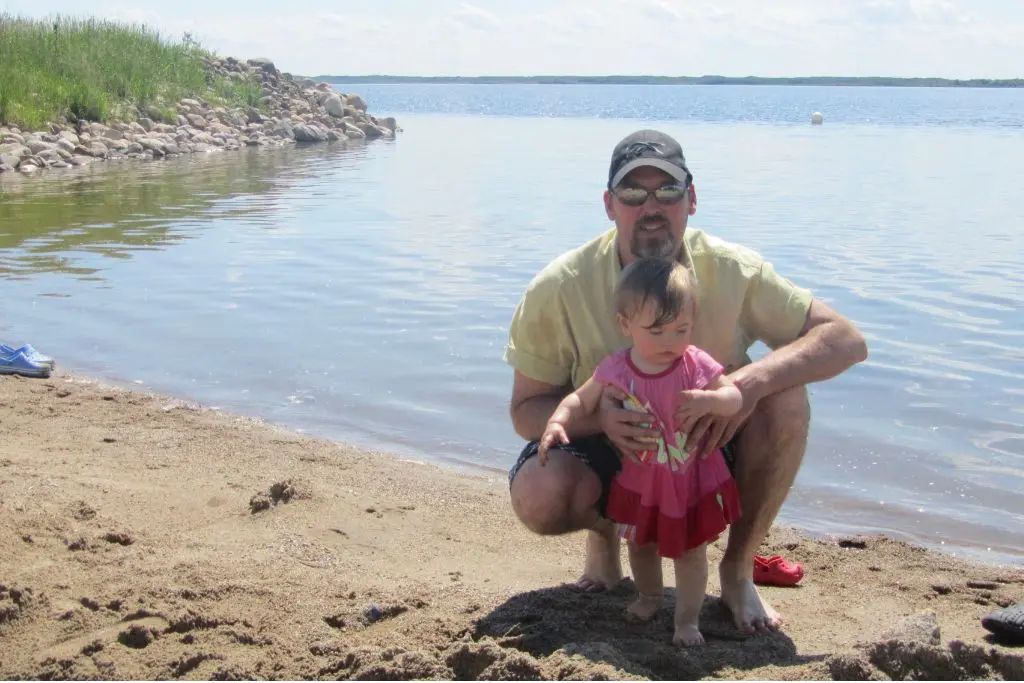 Adult and toddler standing at the sandy shoreline of Douglas Provincial Park with calm lake water in the background.