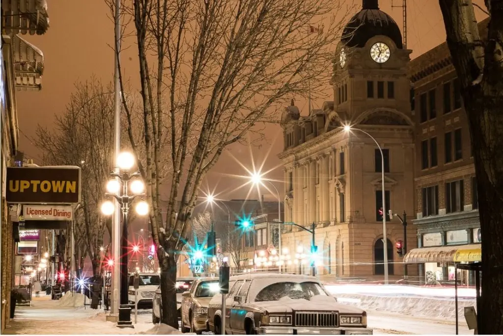 Winter evening street scene in downtown Moose Jaw with snow-covered cars and the historic clock tower lit at night.