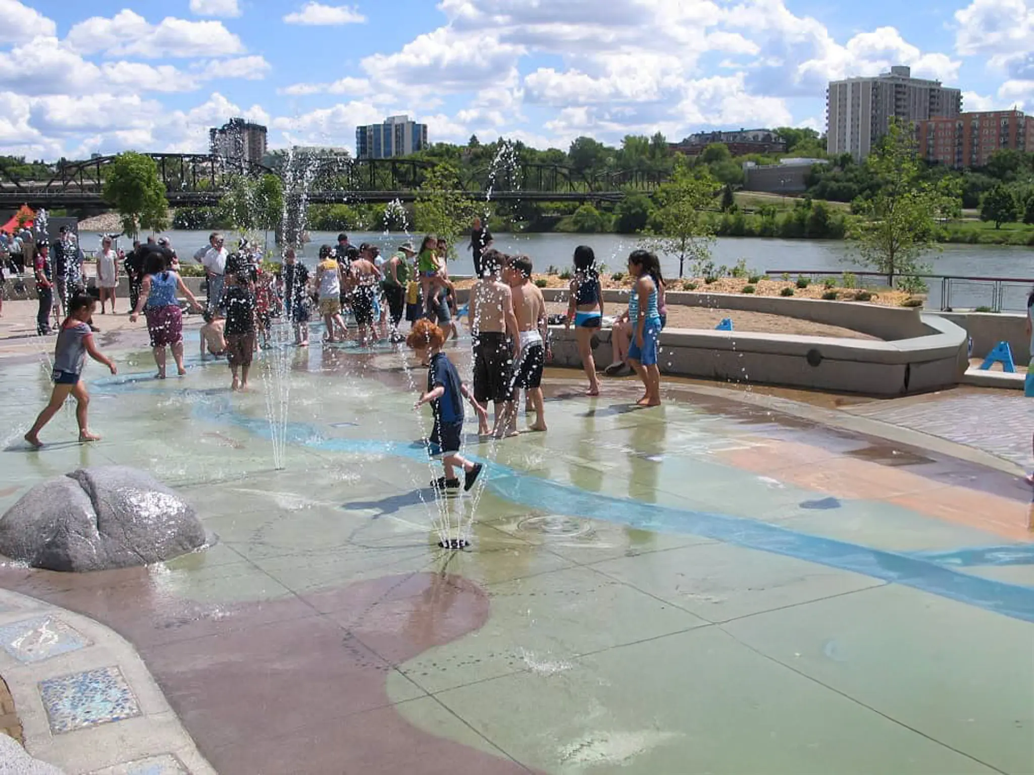 Busy summer scene at River Landing Spray Park with water jets and families cooling off by the river.