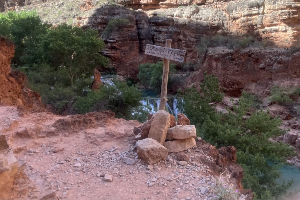 Sign on the trail reading Beaver Falls with slight view of Beaver Falls in the background and canyon walls