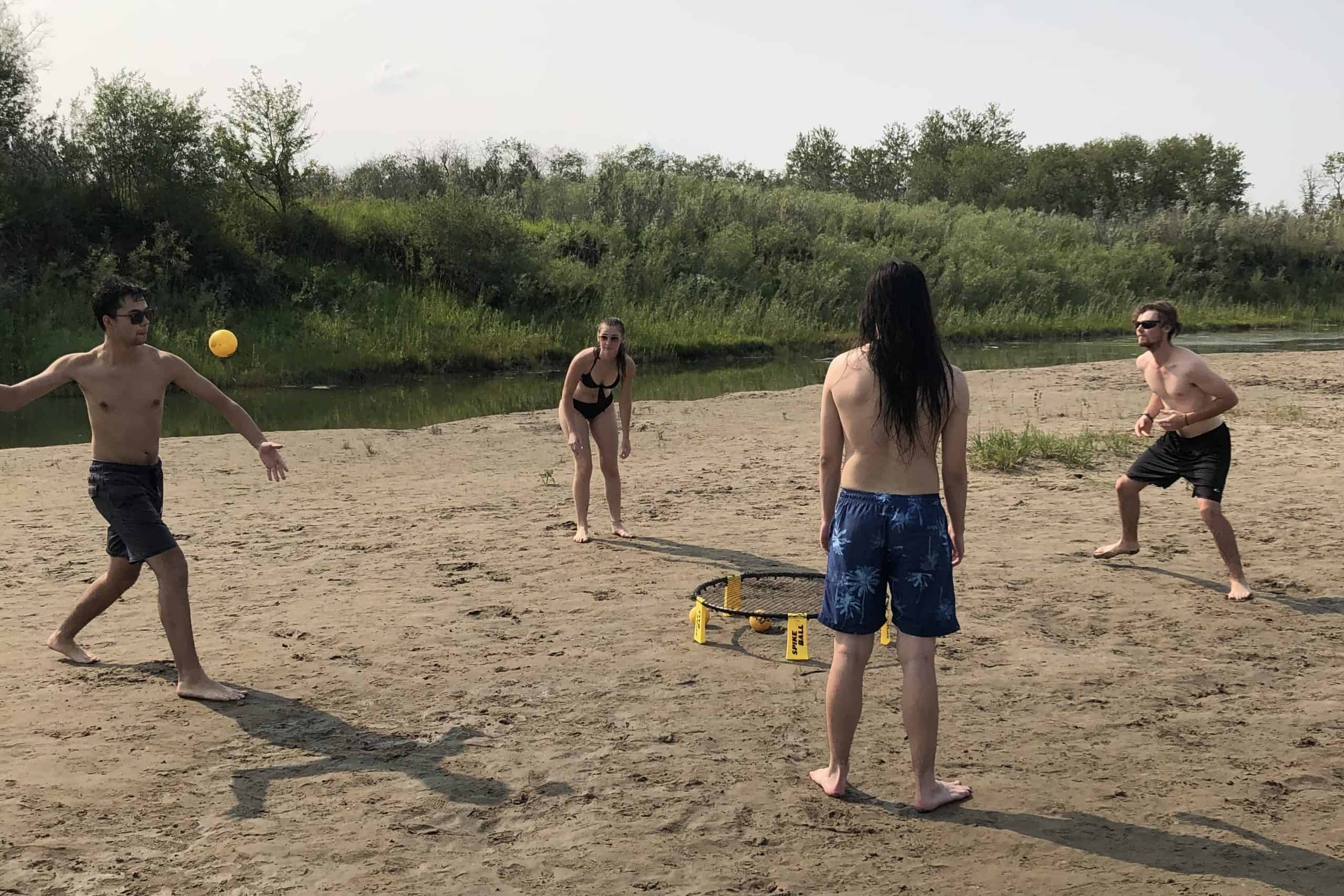 Four people playing spikeball on a sandy riverbank near Poplar Bluffs in Saskatoon during summer.