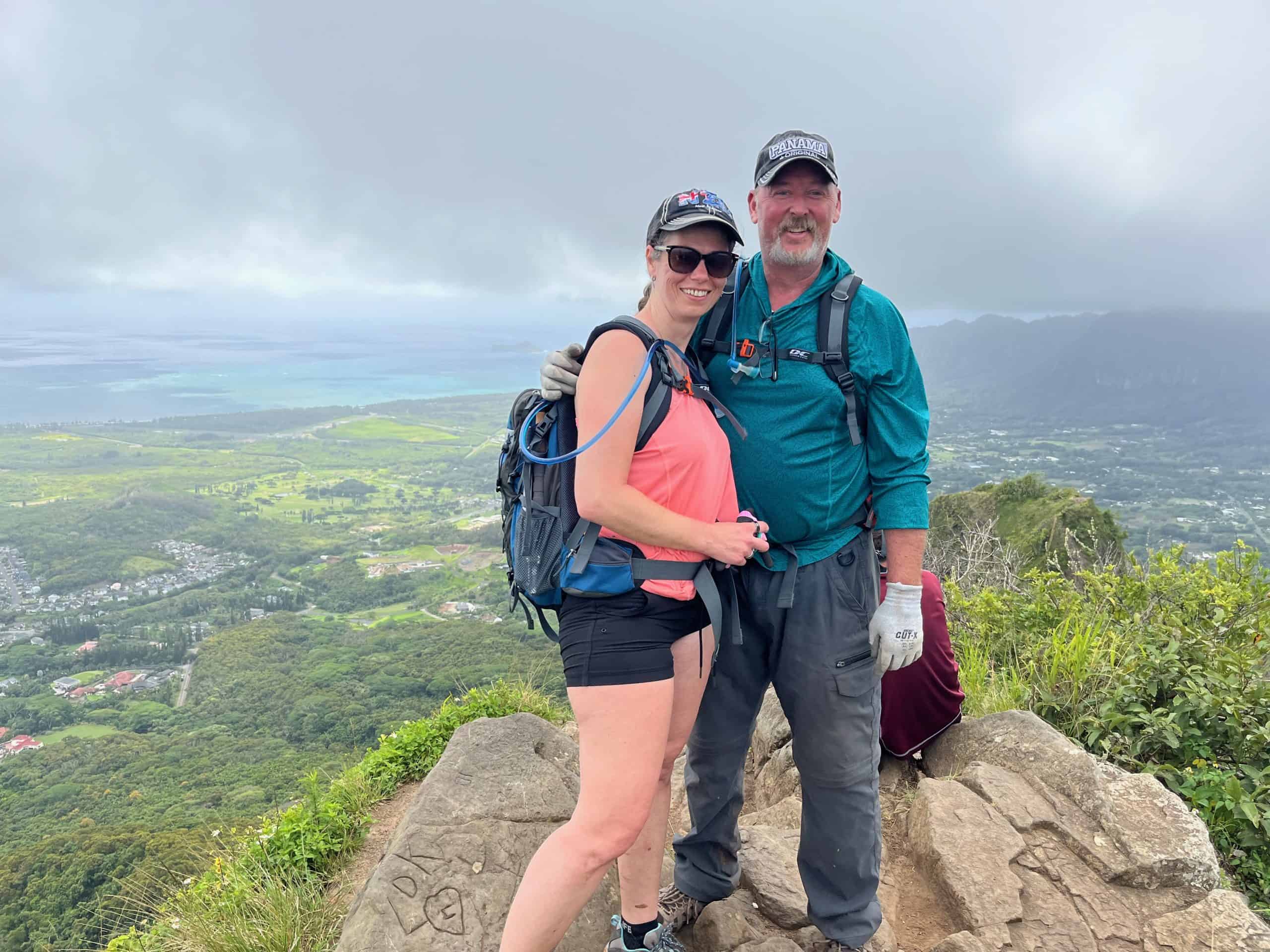 two hikers standing at the top of Peak 1 on Olomana Trail with view of Oahu's Windward Side in background.