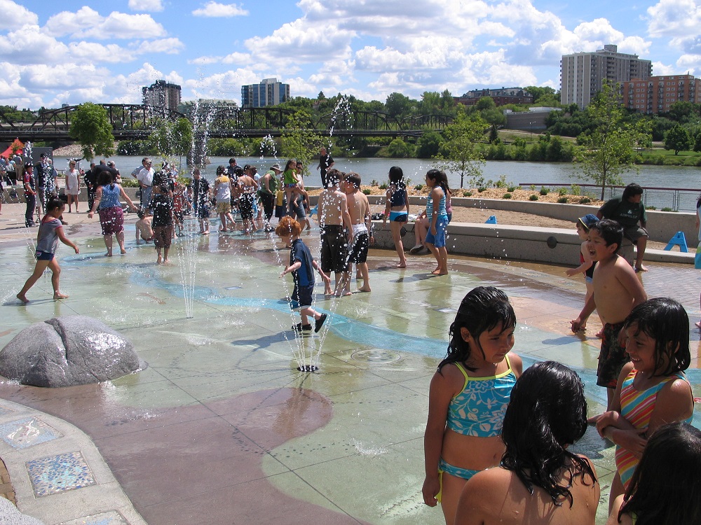Free things to do in saskatoon Children playing at the free River Landing Spray Park with a view of the river and city skyline in the background.