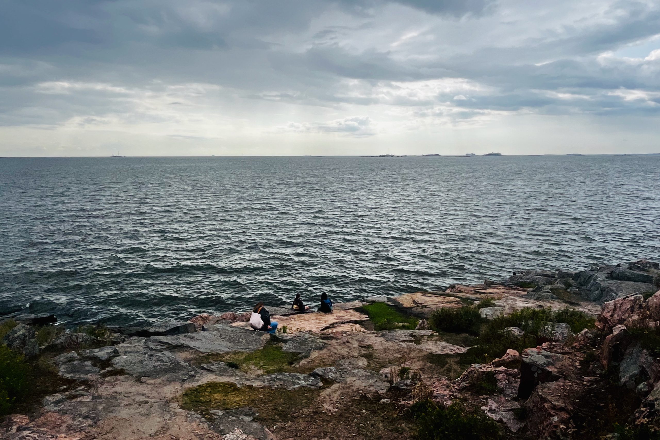 People sitting on the rocky shoreline of Suomenlinna, enjoying views across the Gulf of Finland — a popular day trip destination from Helsinki