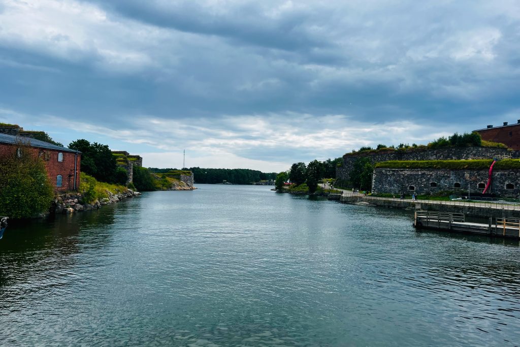 The canal at Suomenlinna fortress in Helsinki, with stone fortifications and historic red-brick buildings along the water under a cloudy sky.