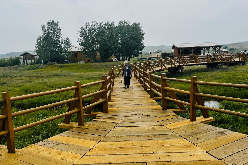 Valley of 1000 Devils Mom walking on the long curved wooden bridge leading the Valley of 1000 Devils trailhead with campground office and cook shelter in the background.