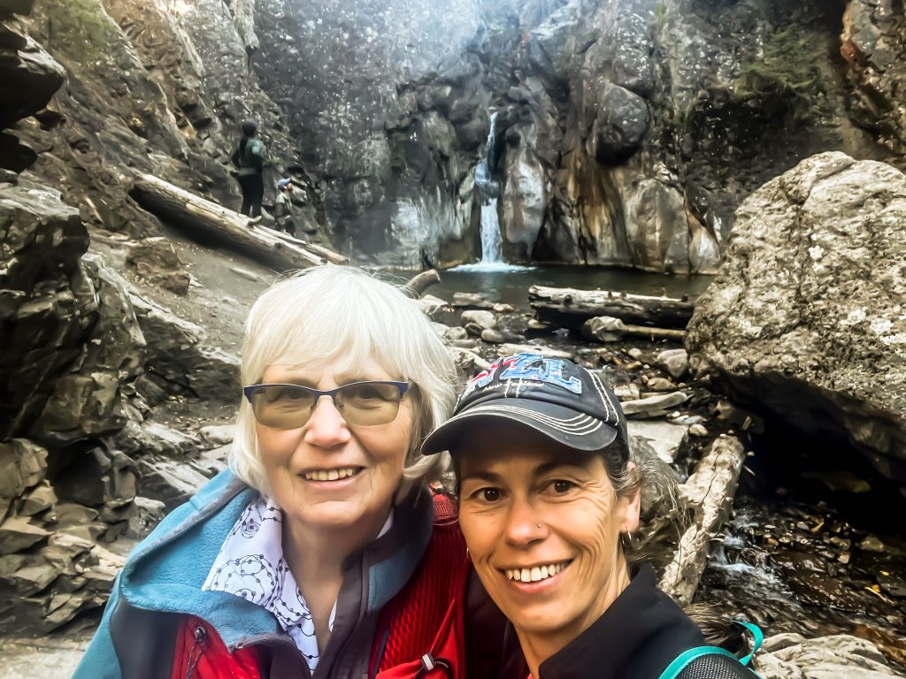 Off the beaten path Kananaskis hikes Two hikers posed in front of Cat Creek Falls wth the falls, boulders, logs, and canyon walls in the background.