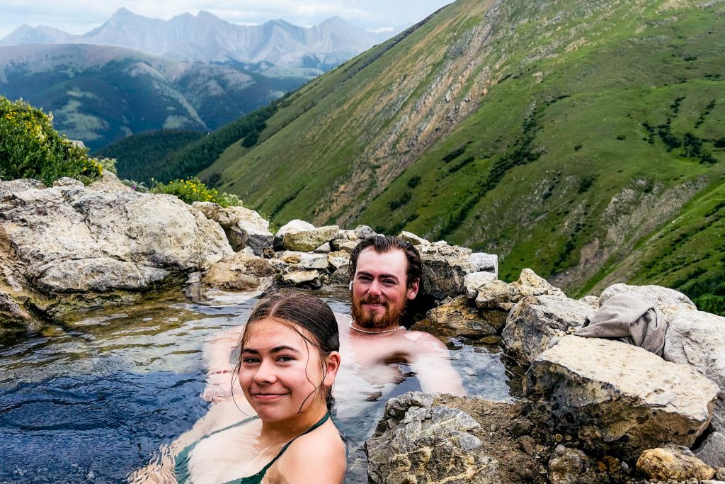 off the beaten path Kananaskis hikes Two hikers relaxing in the natural hot spring pool mid day up Mist Mountain. Mountain views in the background.