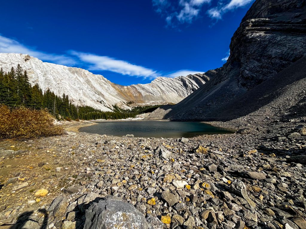 Off the beaten path Kananaskis trails The third Picklejar Lake with rocks in the foreground, mountains in the background, and a dark blue sunny sky.