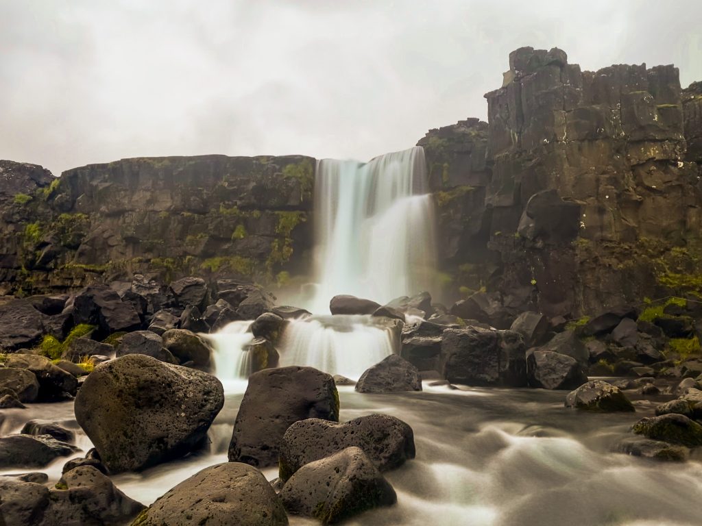Long-exposure view of Oxarárfoss waterfall in Þingvellir, with water flowing over rocks in the foreground and a grey, cloudy sky above.