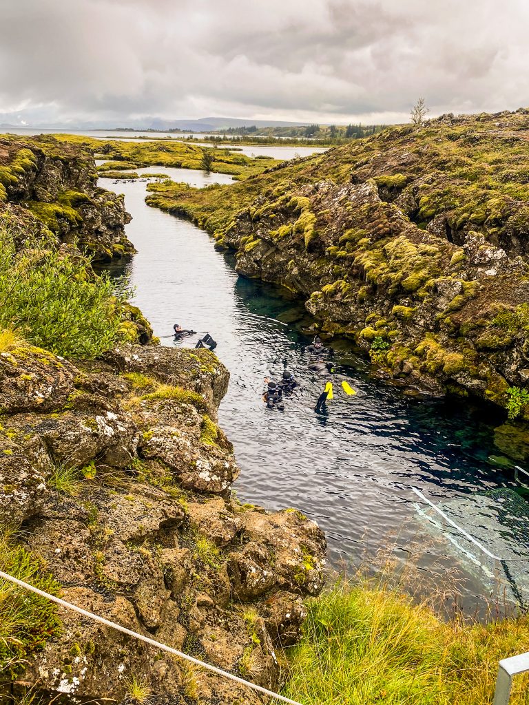 Snorkelers in Silfra Fissure, Þingvellir, seen from above with clear blue water and rocky edges.