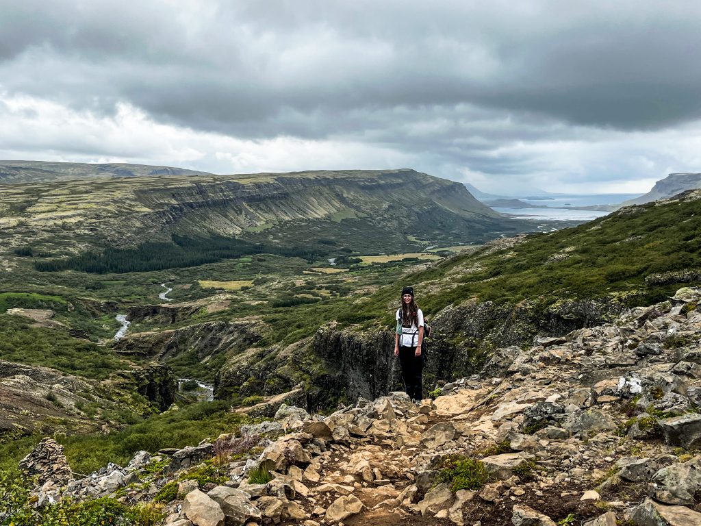 Hiker standing on a rocky slope with a sweeping view of the valley and distant coastline behind on the Glymur trail.