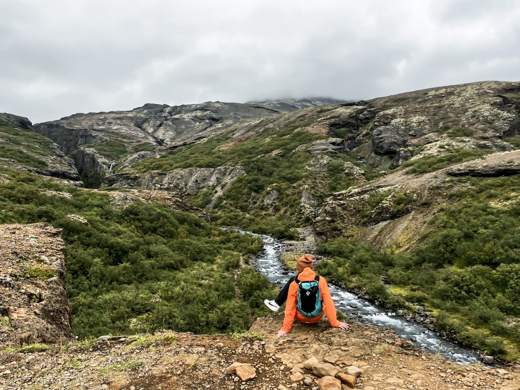 Person sitting on a rocky edge above a river surrounded by green shrubs and mountains during the return portion of the Glymur hike.
