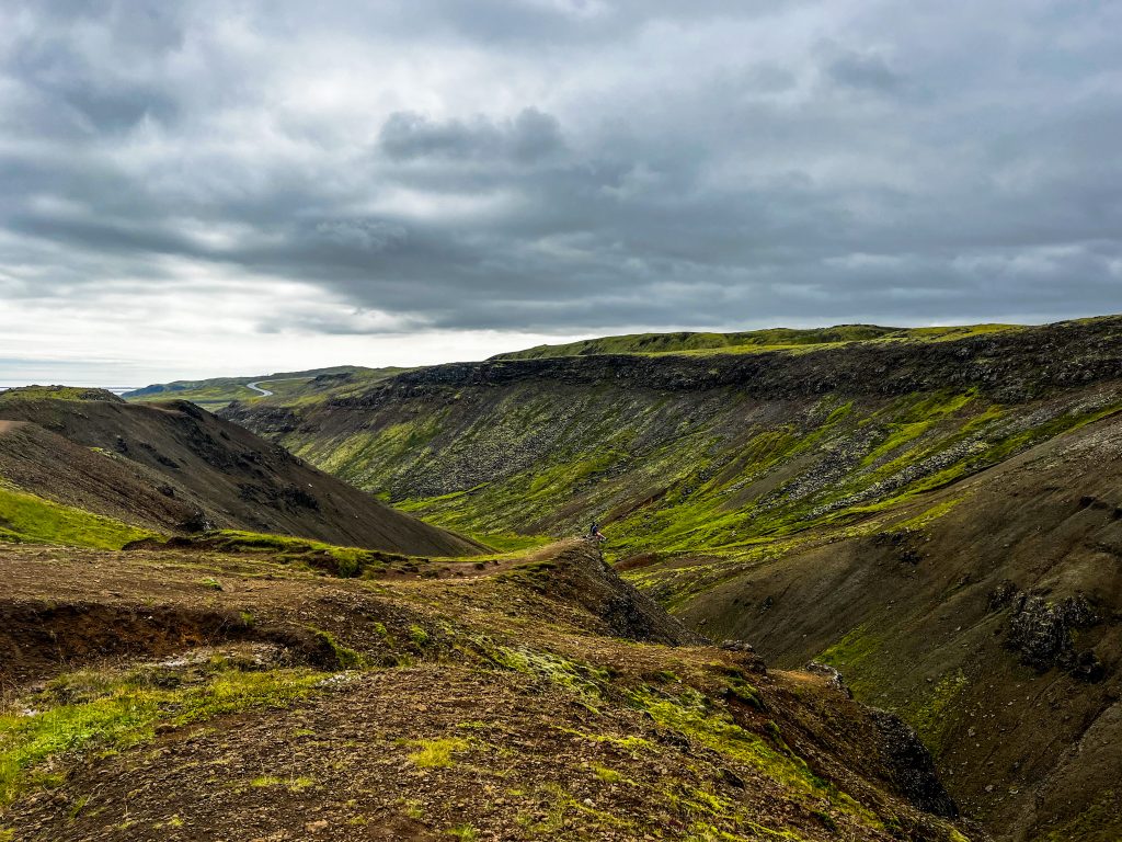 A wide view of the Reykjadalur valley with green mossy slopes, rugged brown hills, and a winding trail under a cloudy sky