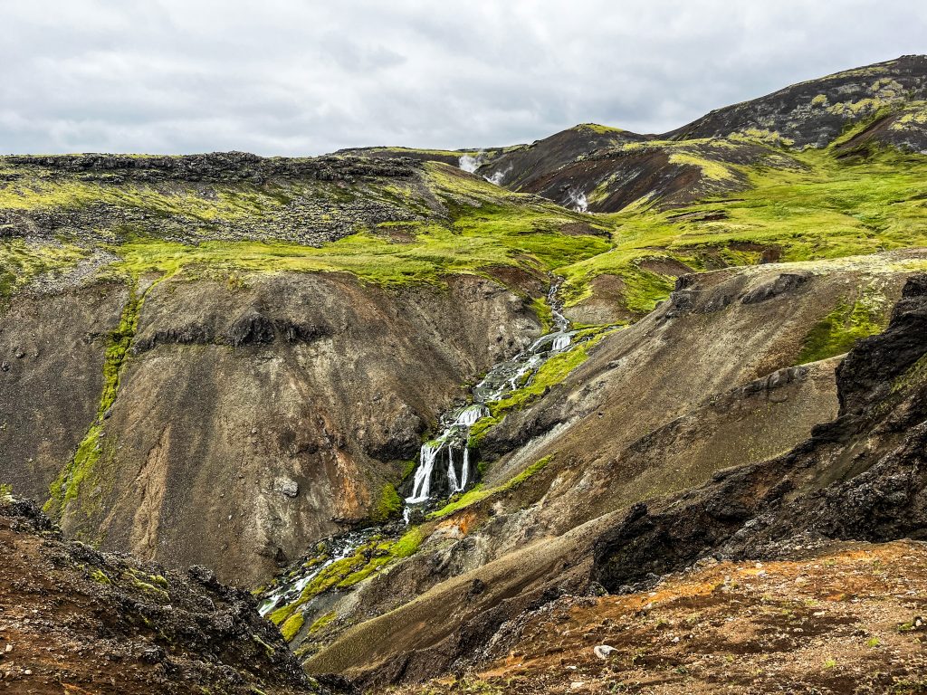 A rocky geothermal hillside with a multi-tiered waterfall running through green moss and mineral-stained slopes.