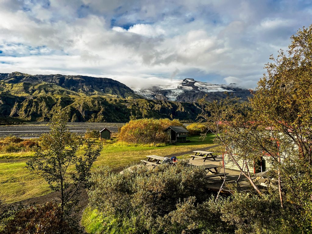 Laugavegur and Fimmvörðuháls combo Picnic tables and small huts at Þórsmörk Langidalur campground, surrounded by birch trees, a wide valley floor, and glacier-covered mountains under a cloudy sky.