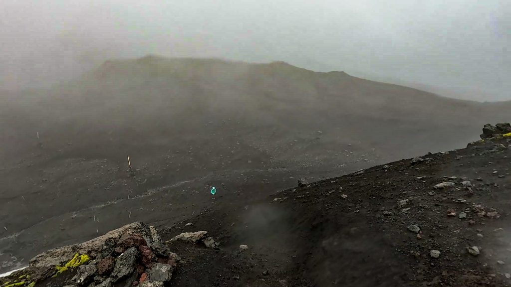 Hiker walking across black volcanic ash and scree on the Fimmvörðuháls trail in foggy, stormy conditions.
