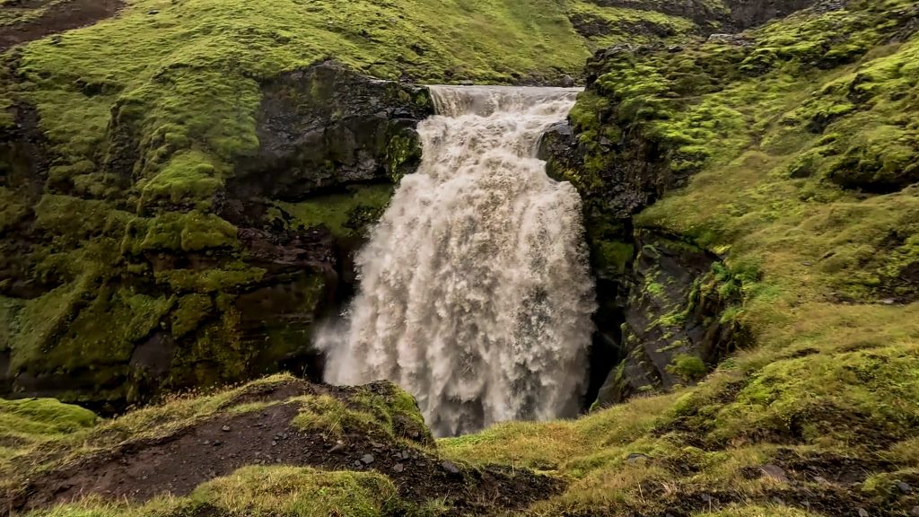 Water rushing over a small, powerful waterfall framed by moss-covered rock and green hills on Iceland's Fimmvörðuháls trail.