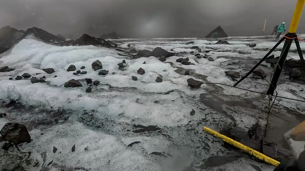 Glacier crossing on the Fimmvörðuháls trail with snow, embedded rocks, and rope-assisted markers in low visibility.