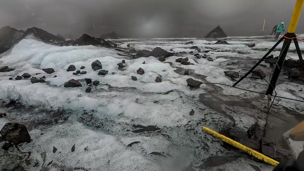 Glacier crossing on the Fimmvörðuháls trail with snow, embedded rocks, and rope-assisted markers in low visibility.