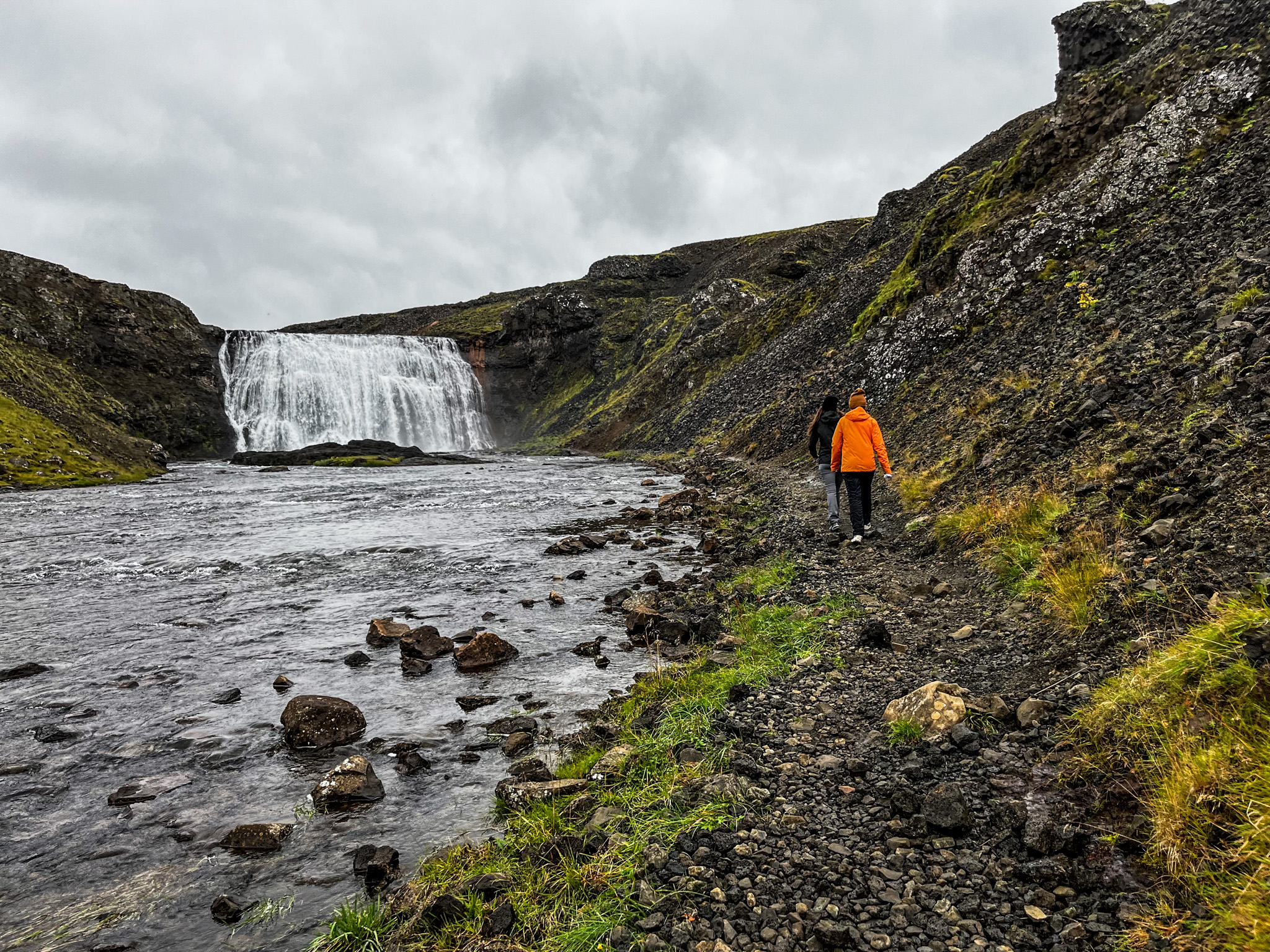 Two people walking on a rocky riverside trail leading toward Þórufoss waterfall in a green, mossy canyon.