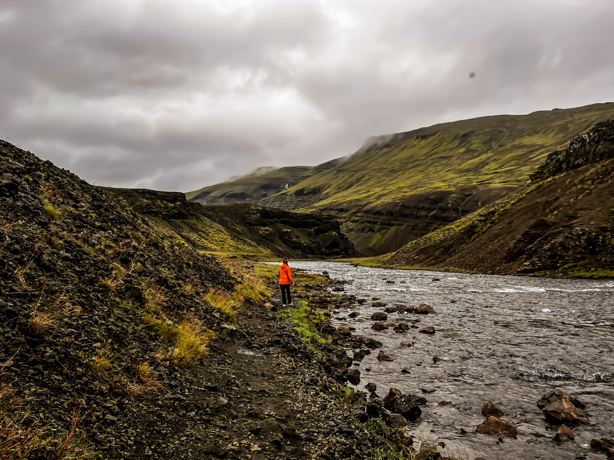 A person walking beside the river through a moss-covered canyon on the trail back from Þórufoss under cloudy skies.