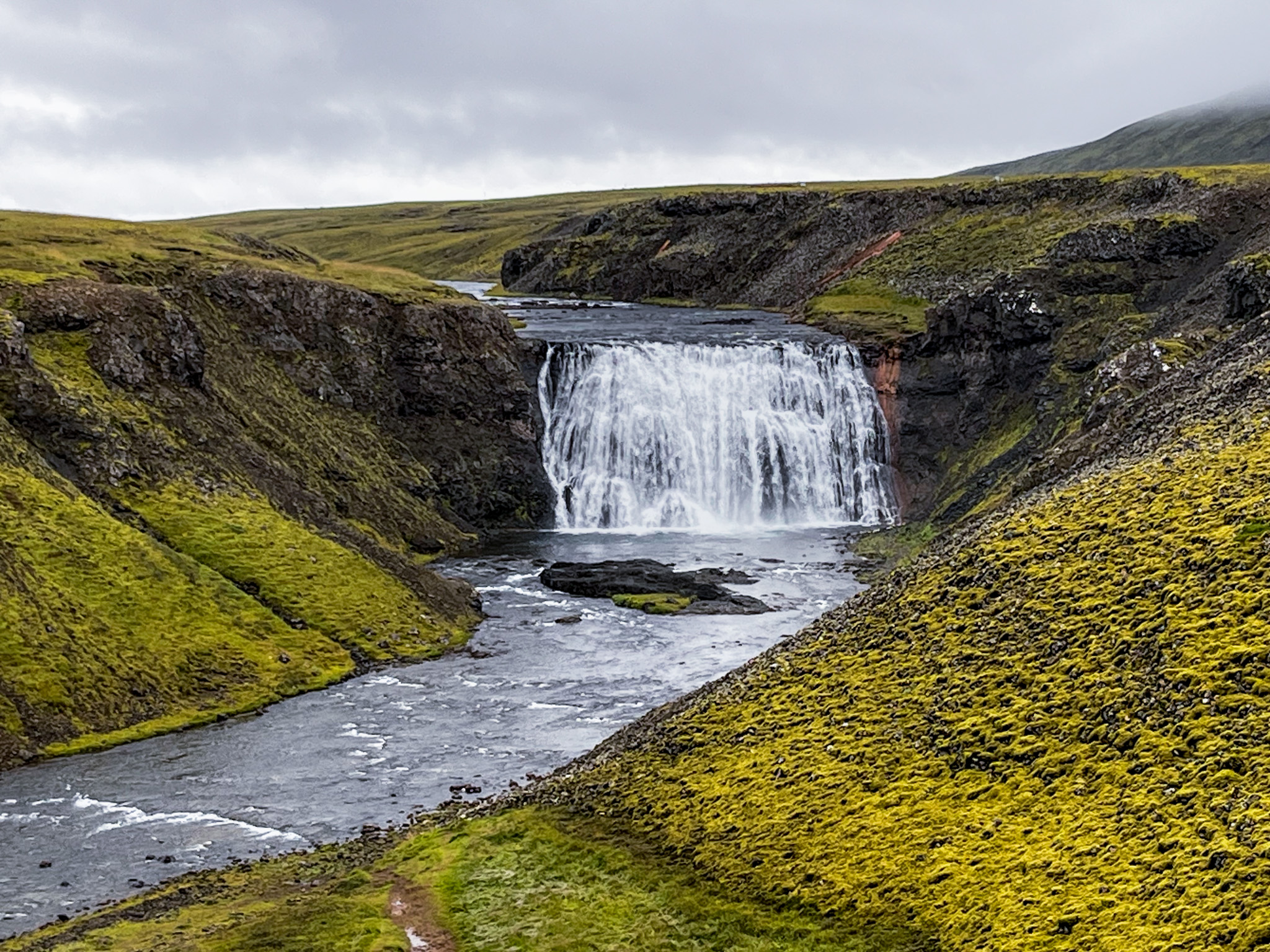 A wide view of Þórufoss waterfall cascading into a moss-covered canyon with a winding river in the foreground.