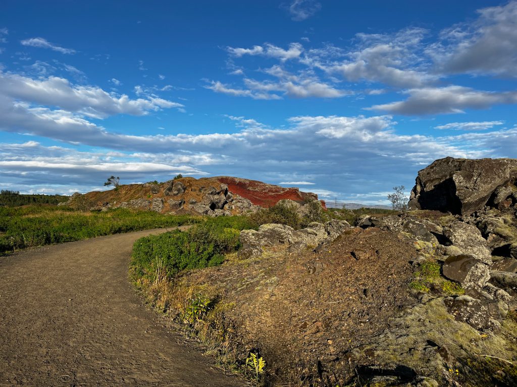 A dirt trail leading toward the red volcanic hills of Rauðhólar under a bright blue sky with scattered clouds.