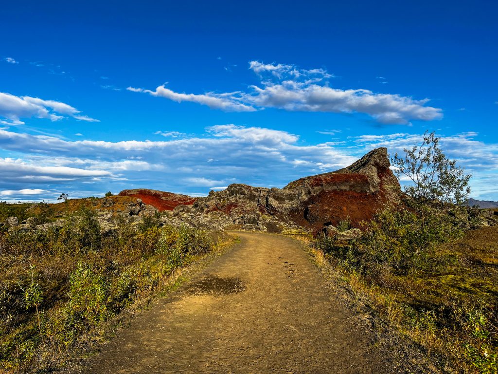 A wide dirt path passing vibrant red volcanic hills and scattered lava rocks at Rauðhólar under a bright blue sky