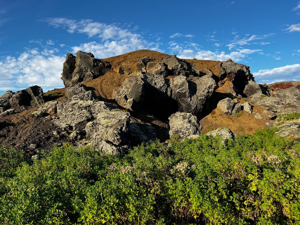 Large dark lava rocks and red volcanic soil at Rauðhólar with green shrubs in the foreground and a blue sky above.