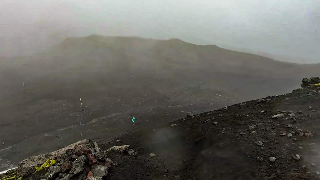 Hiker walking across black volcanic ash and scree on the Fimmvörðuháls trail in foggy, stormy conditions.