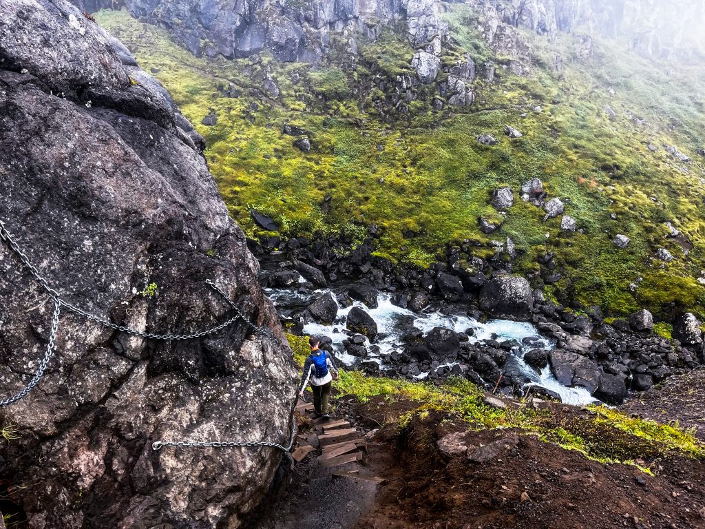Hiker holding the chain on the side of a cliff while descending wooden stairs to get closer to Fardagafoss 