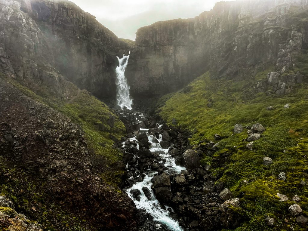 A narrow canyon funnels water from the upper falls into a rocky stream below, framed by steep volcanic walls and mossy slopes.