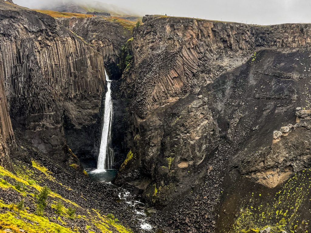 Litlanesfoss waterfall dropping into a narrow basalt canyon, framed by dark rock walls and layered volcanic formations in East Iceland.