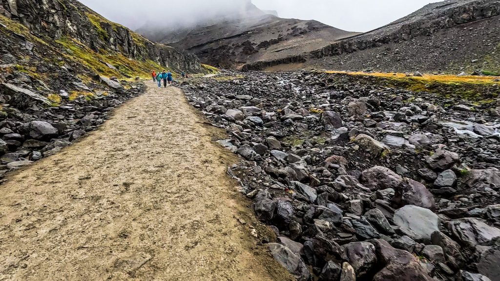 Hikers walking along a gravel trail through a rocky Icelandic valley, with volcanic slopes, loose scree, and low clouds overhead.