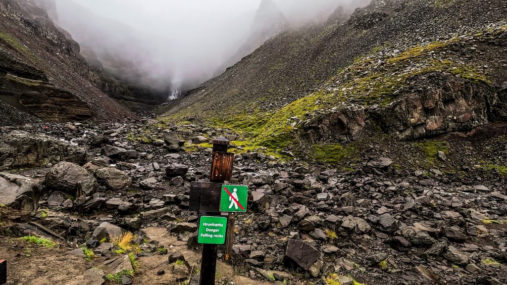 Trail-end warning sign with views of Hengifoss in the distance obscured by fog- loose rocks, steep slopes, and fog filling the narrow valley.
