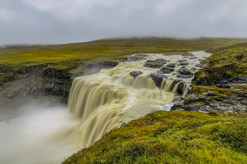 Fax falls in East Iceland roaring over the cliff into a river below white sky overhead and green fields on either side.