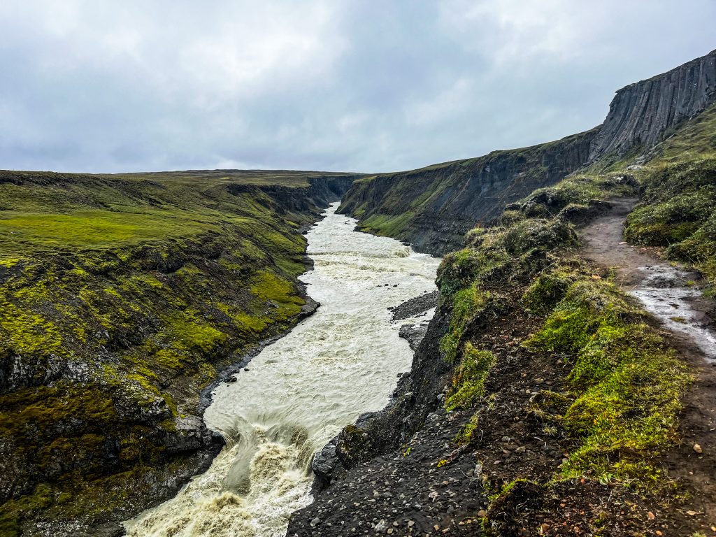 Fast-flowing glacial river cutting through a narrow moss-covered canyon near Laugarfell, with a hiking path following the canyon rim.