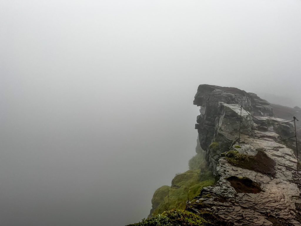 Fog-shrouded cliff edge along the trail at Múlagljúfur Canyon, with a narrow path and rocky outcrop fading into mist.