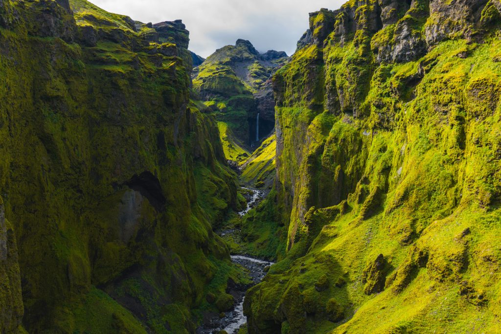 Towering Cliffs of  Múlagljúfur Canyon rising above the river with a distant waterfall in the background
