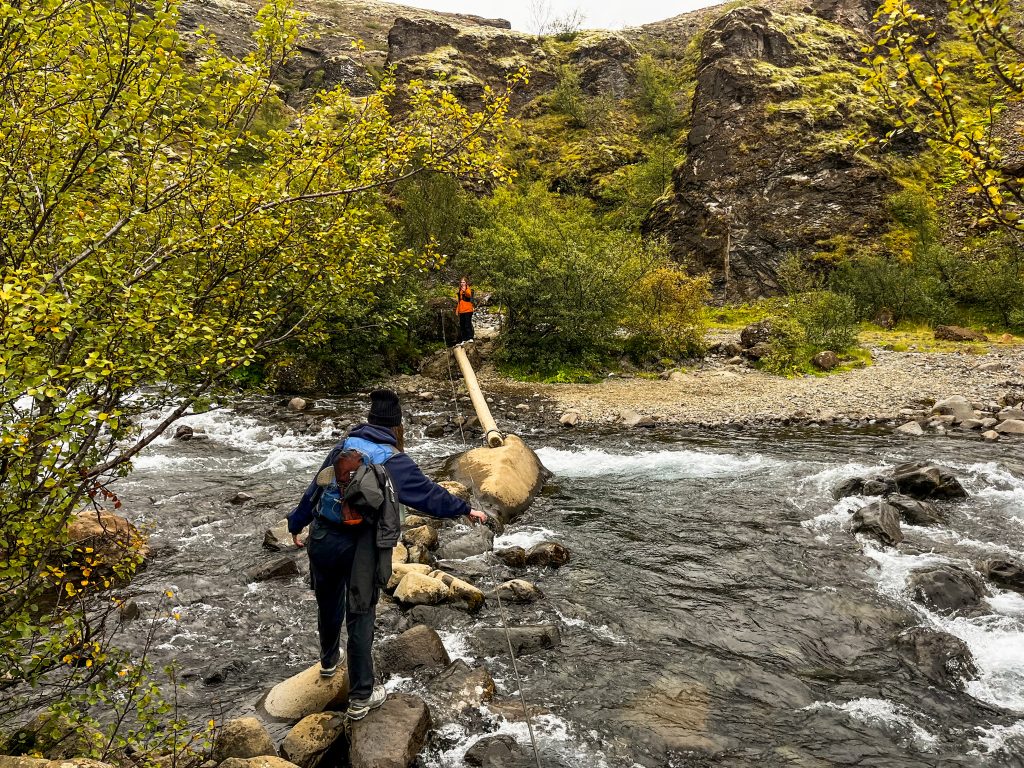 Hikers carefully crossing a shallow river on stepping stones and a narrow log bridge along the Glymur waterfall trail in Iceland