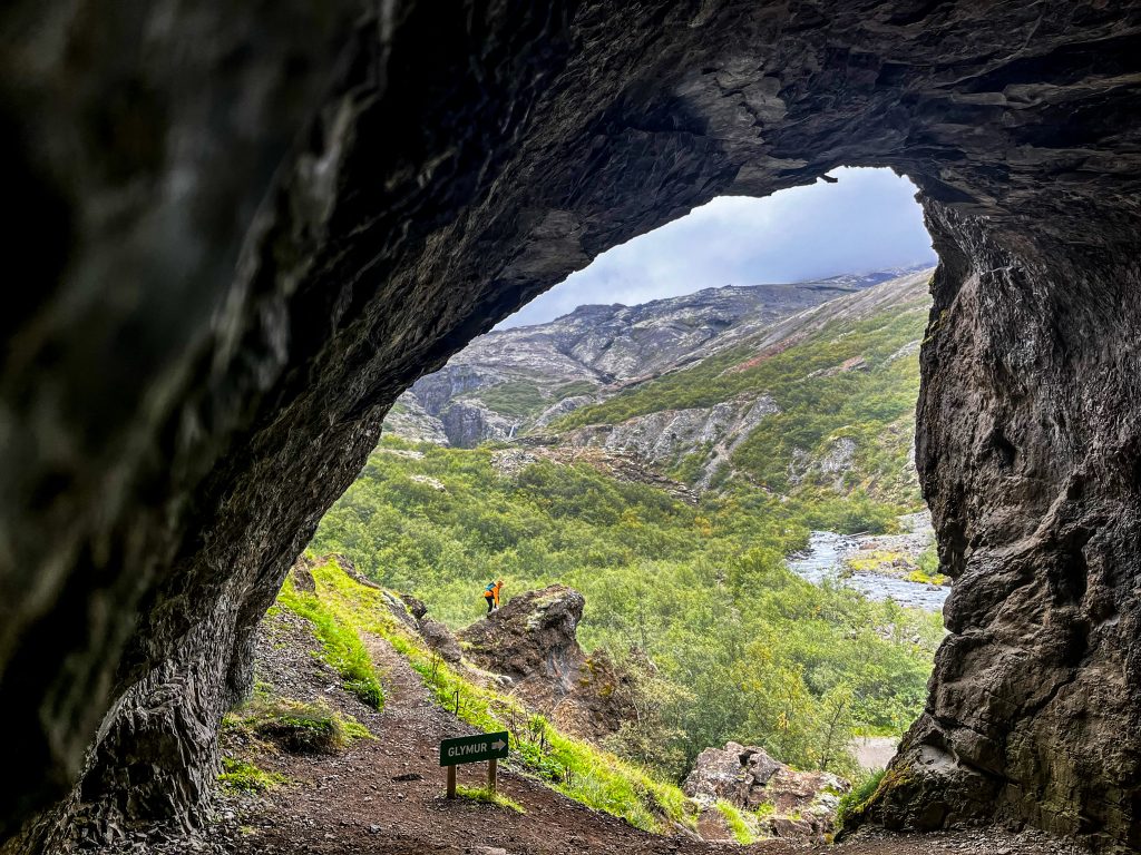 View through a rocky cave opening on the Glymur trail, looking out toward a green valley with a river below and a hiker standing on a cliff edge