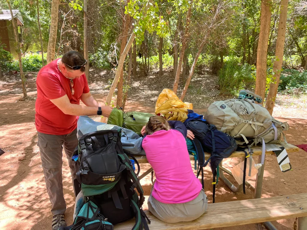 A hiker sitting with her head down on a picnec table surrounded by backpacks while another hiker looks for something in one of the backpacks.