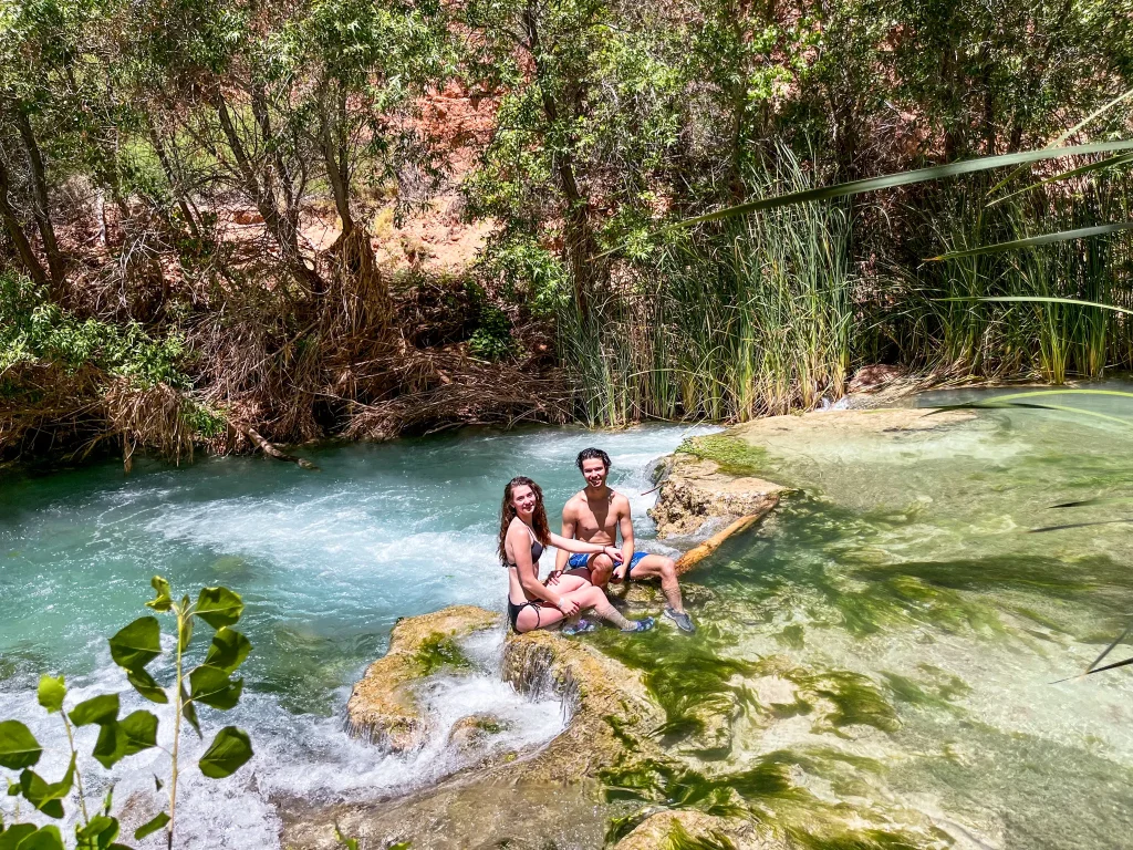 Two people sitting on a small rock ledge in Havasu Creek, surrounded by turquoise water, reeds, and canyon vegetation, cooling off in a natural pool