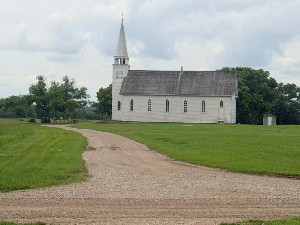 Historic white church at Batoche National Historic Site in Saskatchewan with a gravel road leading to the building across a grassy field.