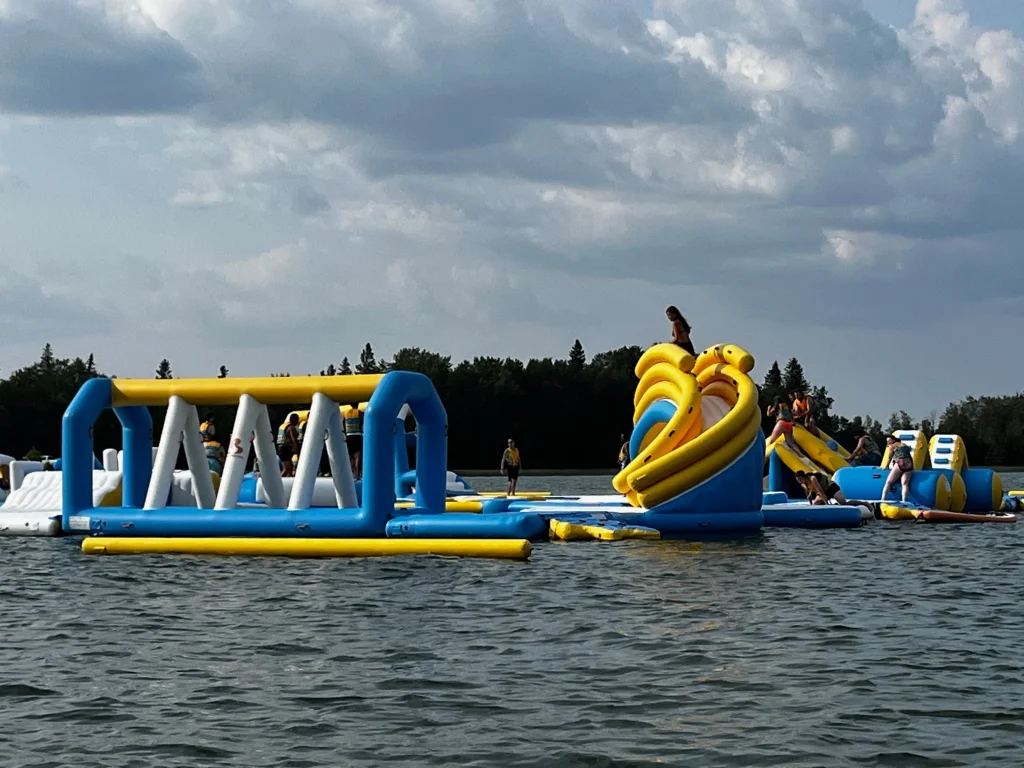 Inflatable water park floating on Candle Lake at Sandy Bay with slides, climbing obstacles, and children playing on the structures.